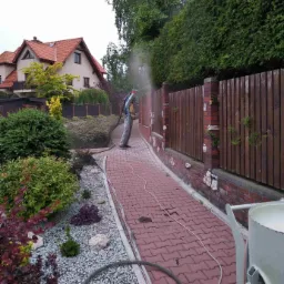 Pracownik w kombinezonie ochronnym czyści ceglany murek z drewnianym płotem za pomocą strumienia ściernego; widoczny wąż i zbiornik urządzenia, kostka brukowa i bujna roślinność w tle.