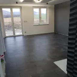 Wnętrze pokoju po remoncie z ciemną podłogą z płytek, szarą ścianą, białymi ścianami z oknami i dekoracyjnym sufitem podwieszanym z oświetleniem LED.