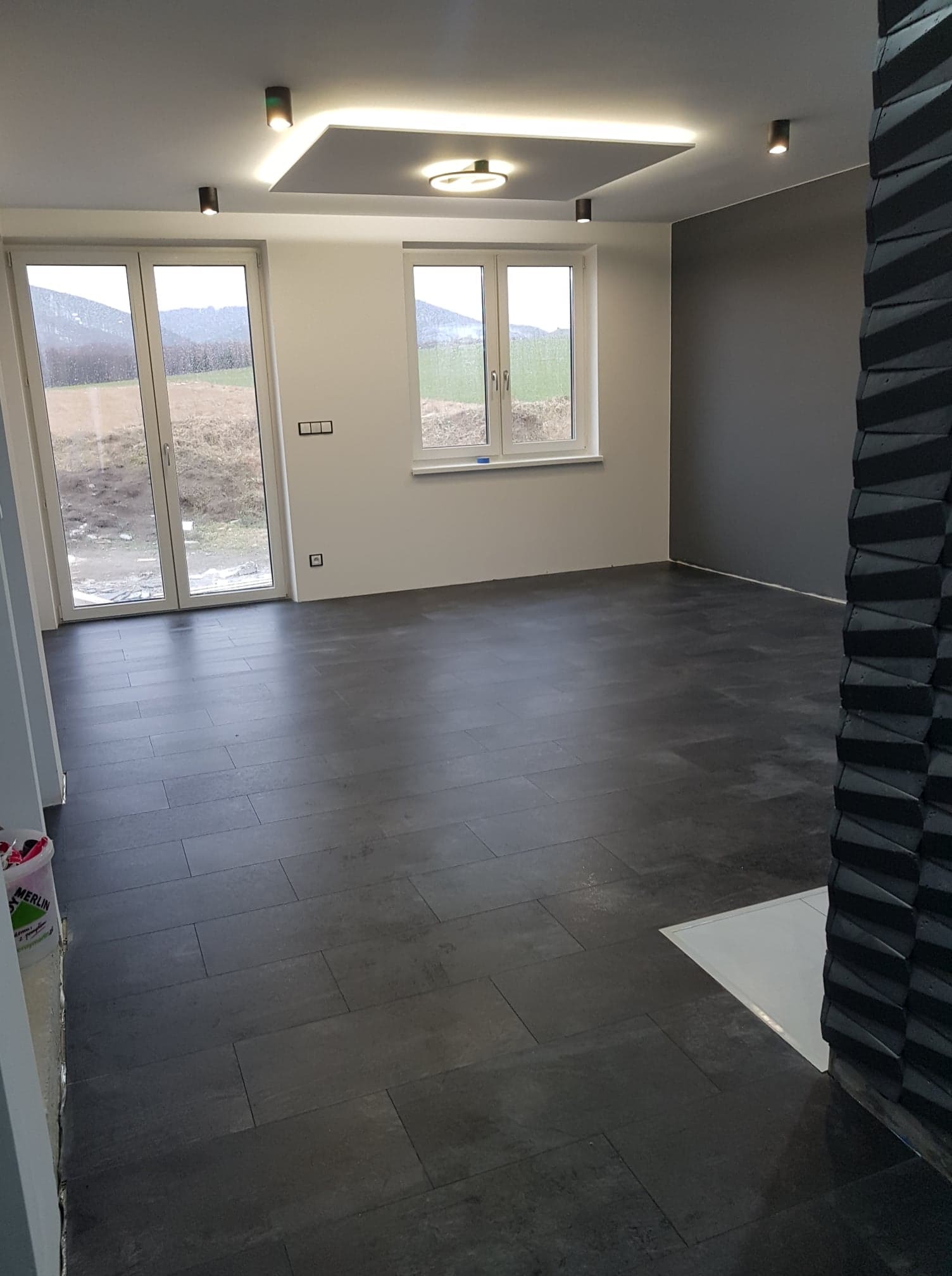 Wnętrze pokoju po remoncie z ciemną podłogą z płytek, szarą ścianą, białymi ścianami z oknami i dekoracyjnym sufitem podwieszanym z oświetleniem LED.