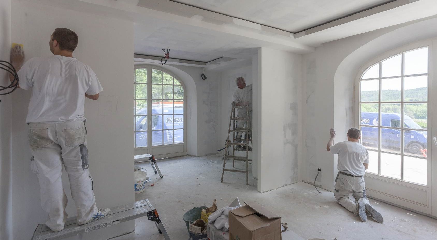 Wnętrze w trakcie przygotowania do malowania: trzech mężczyzn w białych ubraniach, jeden szlifuje ścianę na podwyższeniu, drugi stoi na drabinie, trzeci szlifuje ścianę przy oknie, widoczne...