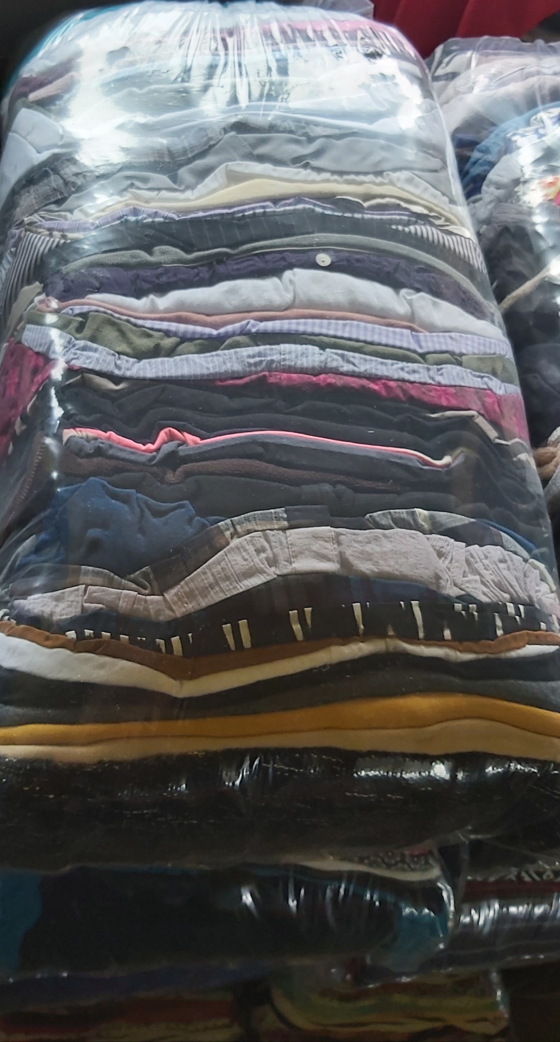 Duży pakiet sprasowanych, używanych ubrań w przezroczystej folii, z widocznymi warstwami różnych tkanin i kolorów, ułożonych jeden na drugim.