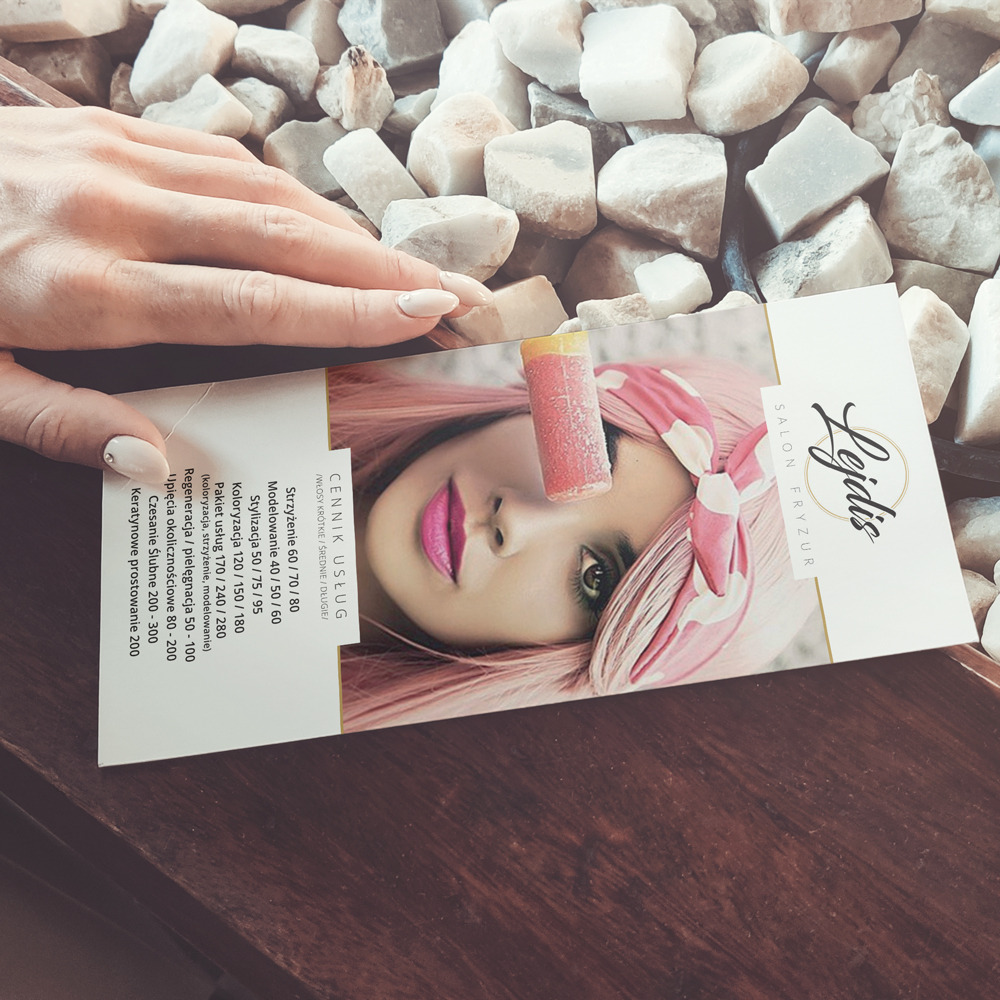 Ulotka salonu fryzjerskiego Lejdis z cennikiem usług, trzymana w dłoni, na tle białych kamieni dekoracyjnych i drewnianego blatu.