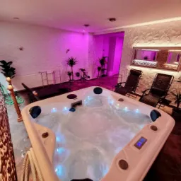 Wnętrze pokoju z jacuzzi wypełnionym wodą, podświetlonym na niebiesko, z dwoma leżakami, roślinami doniczkowymi, lustrem i dekoracyjną ścianą w tle, oświetlone fioletowym światłem.