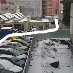 Budynek w trakcie budowy w zimowej scenerii, widoczne fundamenty zabezpieczone folią, obok parking zasypany śniegiem.