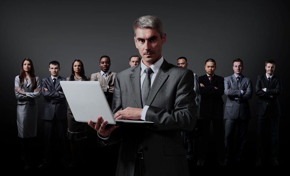 Zespół biznesowy z liderem trzymającym laptopa na tle ciemnoszarego tła.
