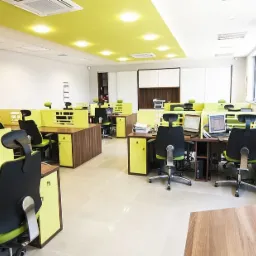 Jasne biuro z rzędami biurek z przegrodami w odcieniach drewna i limonki, czarnymi krzesłami biurowymi i komputerami.