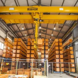 Magazyn z pomalowanymi na pomarańczowo elementami konstrukcyjnymi, transportowanymi przez suwnicę BUDTOR
