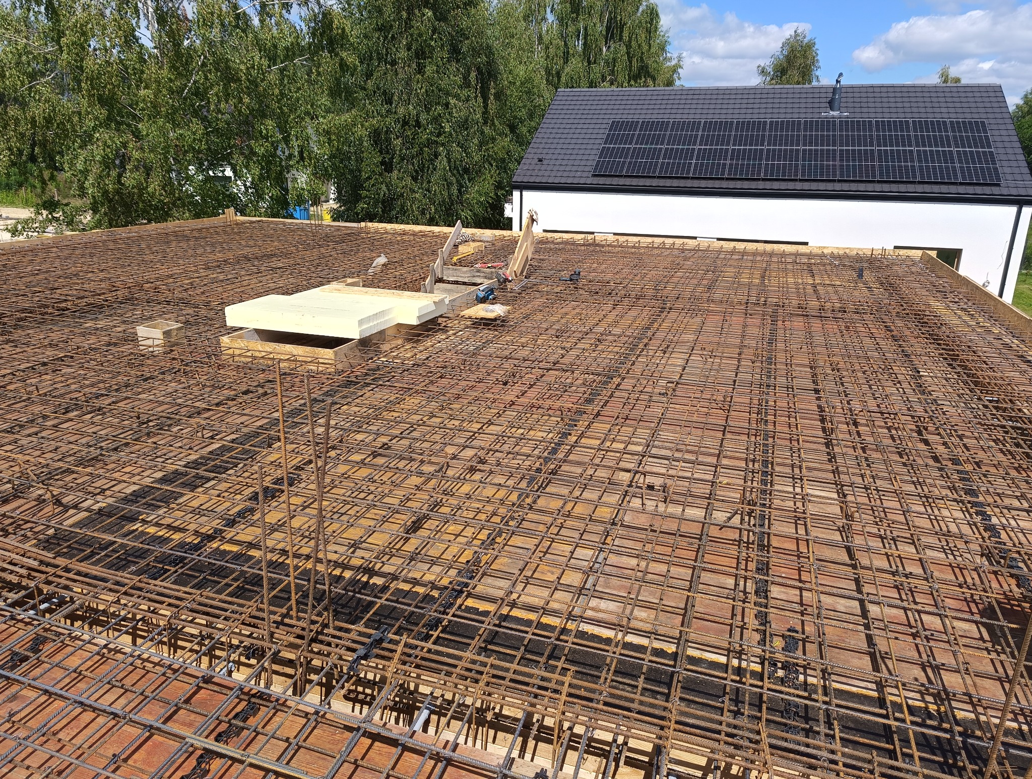 Zbrojenie płyty fundamentowej na drewnianym szalunku, z widocznymi otworami na instalacje i narzędziami budowlanymi w tle, z nowym domem z panelami słonecznymi w tle.