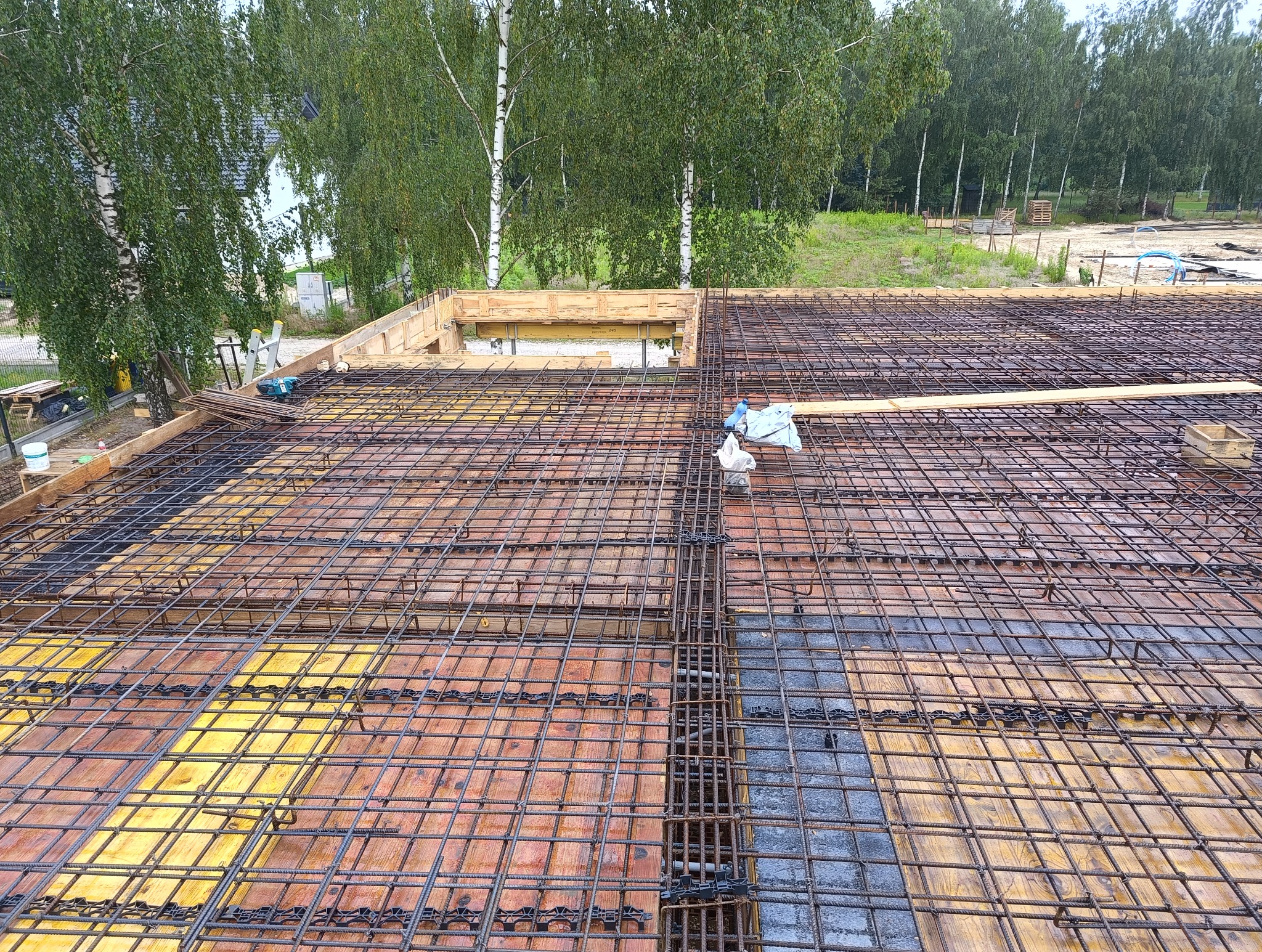 Zbrojenie płyty fundamentowej domu jednorodzinnego z widocznymi deskami szalunkowymi, siatką zbrojeniową i elementami dystansowymi w trakcie budowy, z zielonymi drzewami w tle.