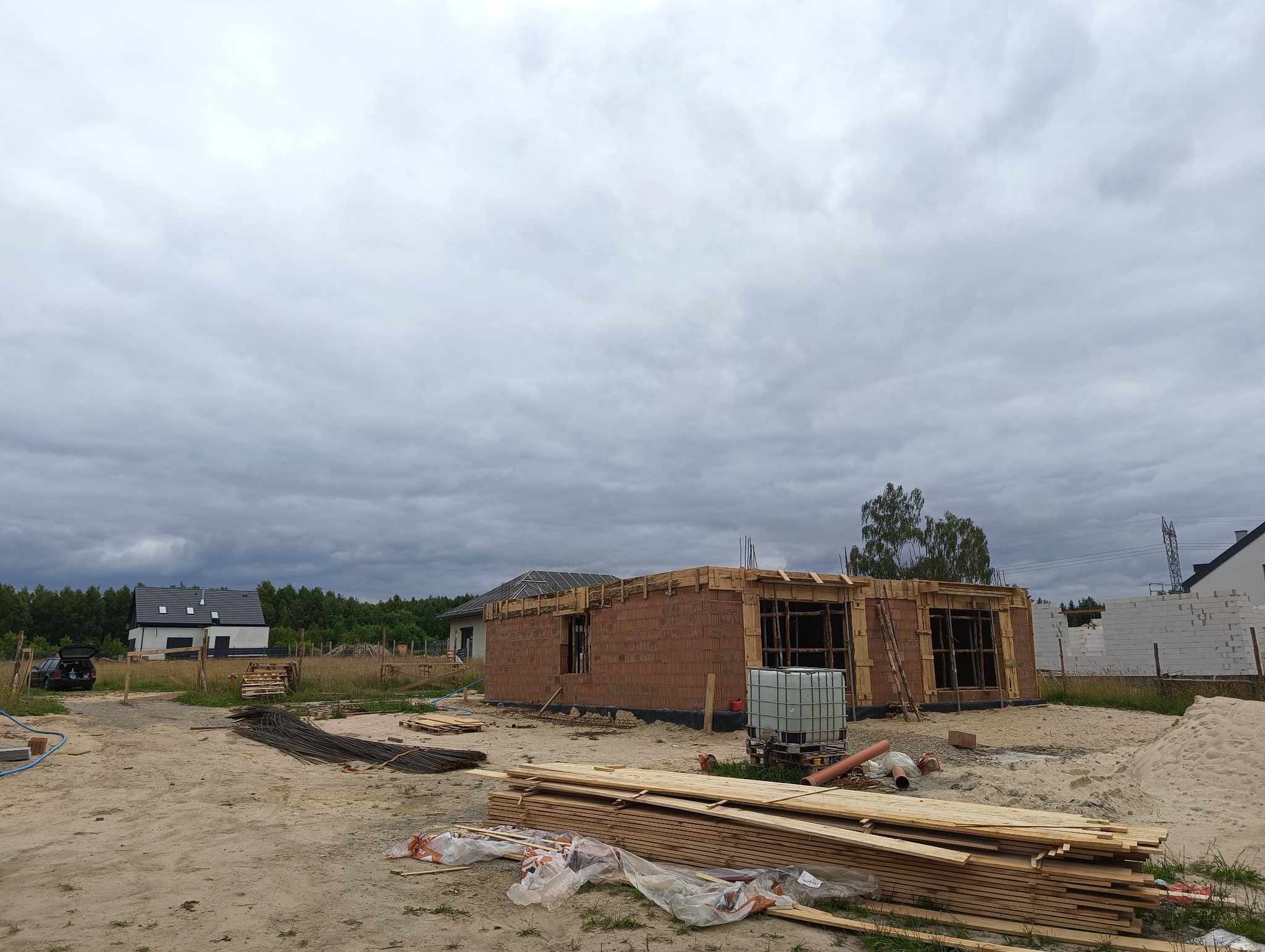 Budowa domu jednorodzinnego w trakcie wznoszenia ścian z czerwonej cegły, widoczne drewniane szalunki okienne i drzwiowe, na pierwszym planie stos desek i materiały budowlane, pochmurne niebo w tle.