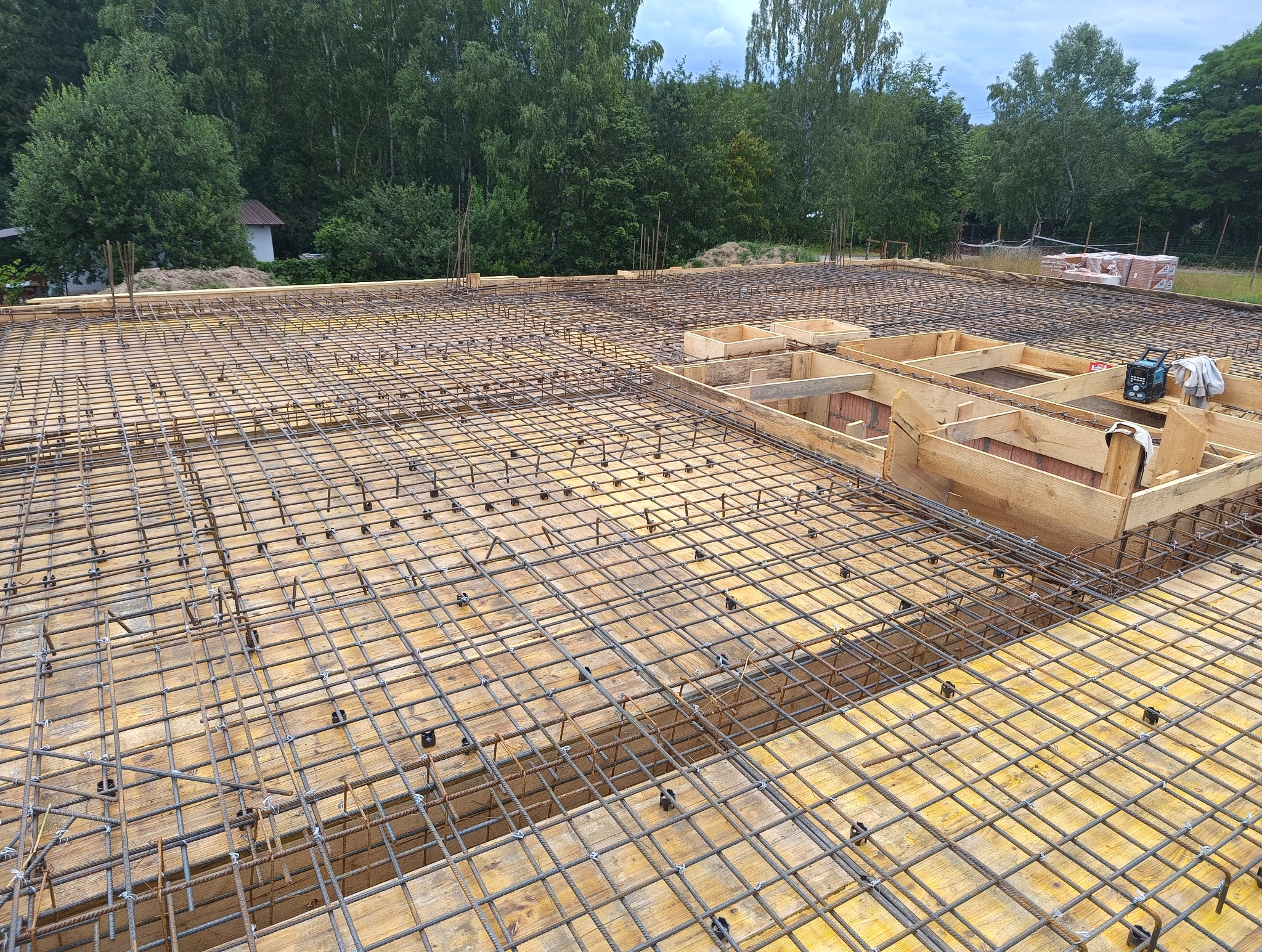 Przygotowanie zbrojenia i szalunku pod wylewkę stropu na budowie domu, widoczne drewniane formy i siatka z prętów stalowych spiętych plastikowymi klipsami, w tle zieleń drzew.