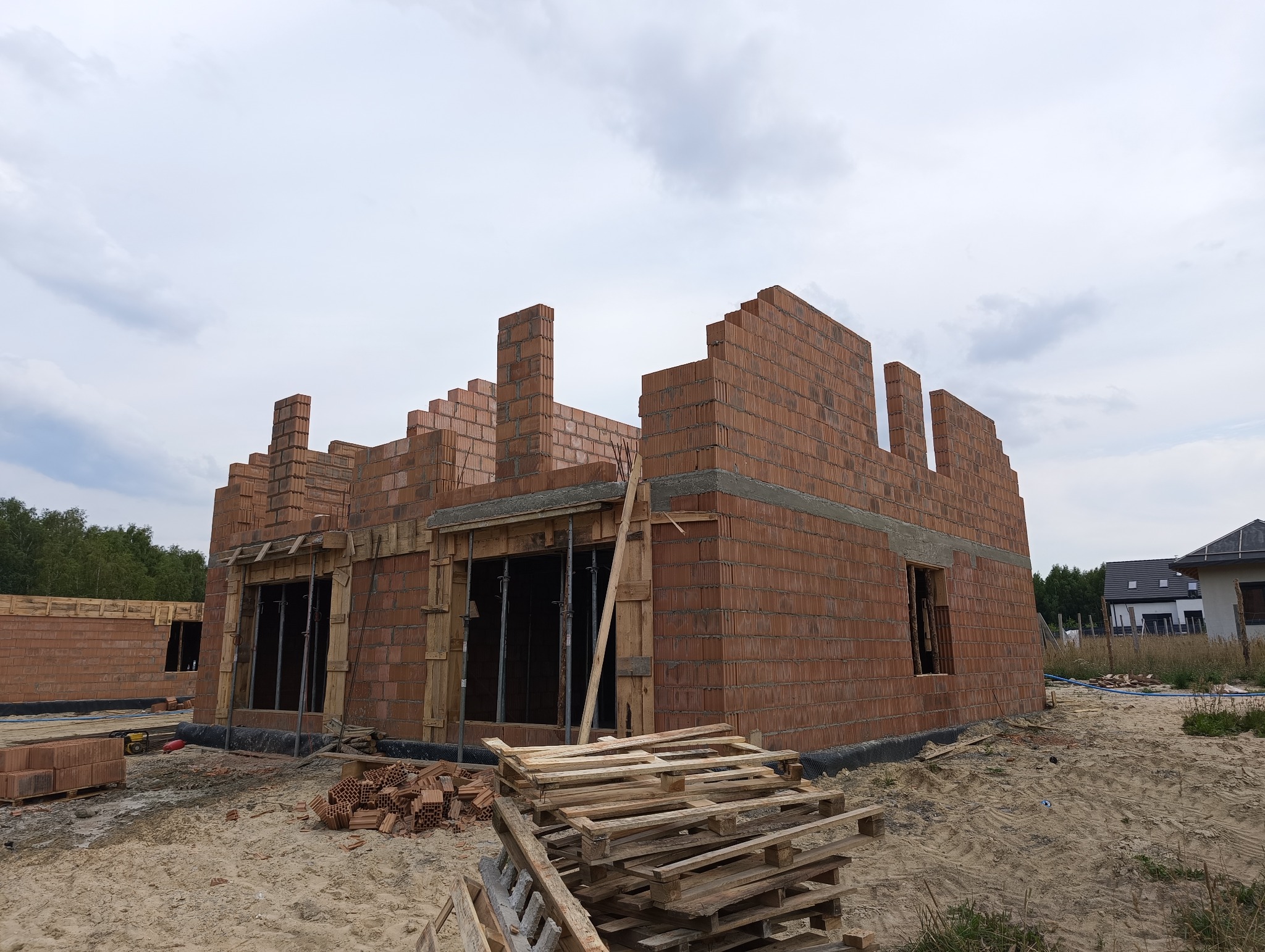 Surowy stan domu jednorodzinnego z czerwonej cegły, widoczne drewniane szalunki okienne, palety i porozrzucane cegły na piaszczystym placu budowy w pochmurny dzień.