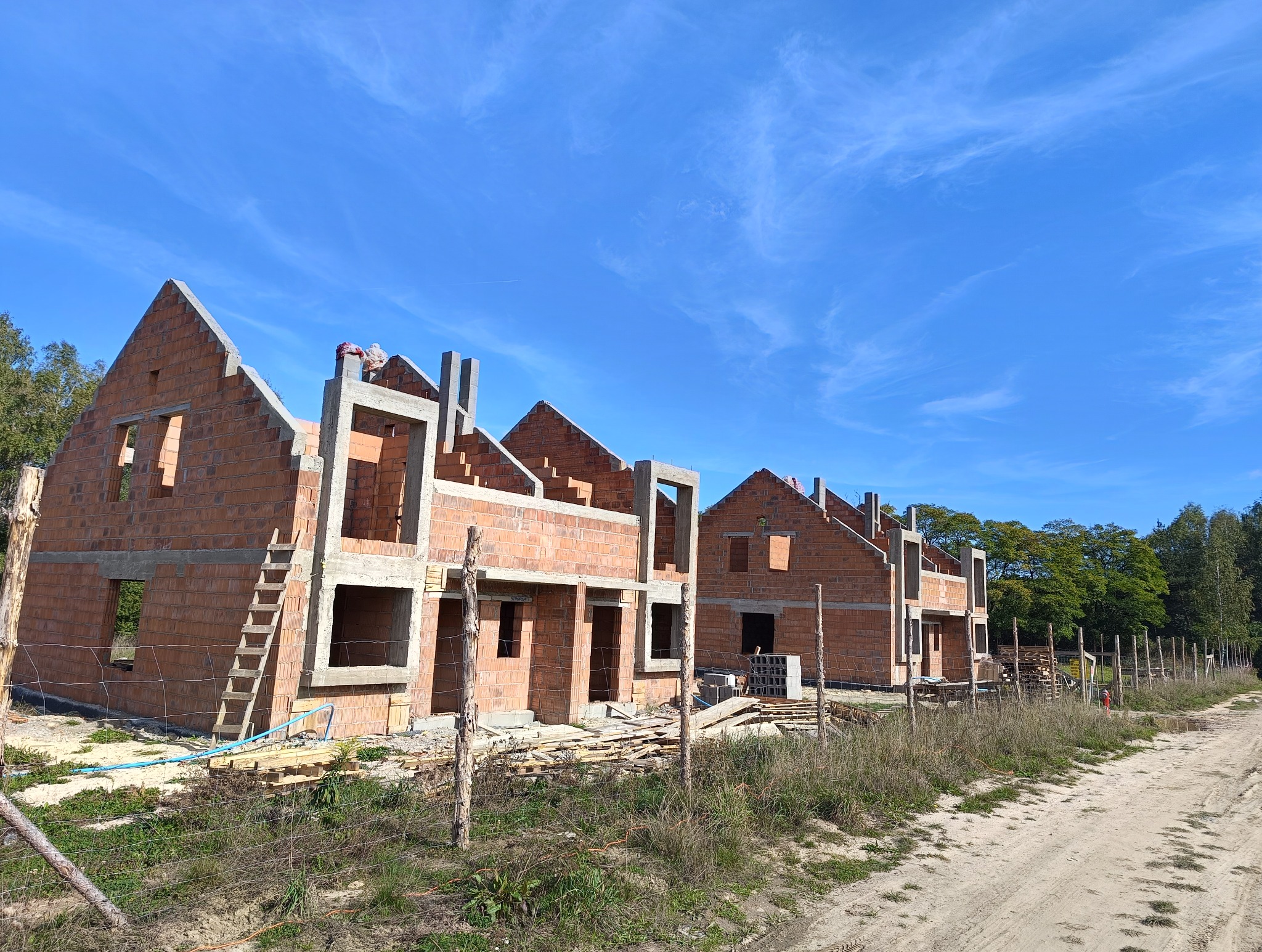 Wczesny etap budowy szeregu domów z czerwonej cegły, widoczne niezadaszone ściany, drewniane rusztowanie oraz drabina oparta o jedną ze ścian, błękitne niebo w tle.