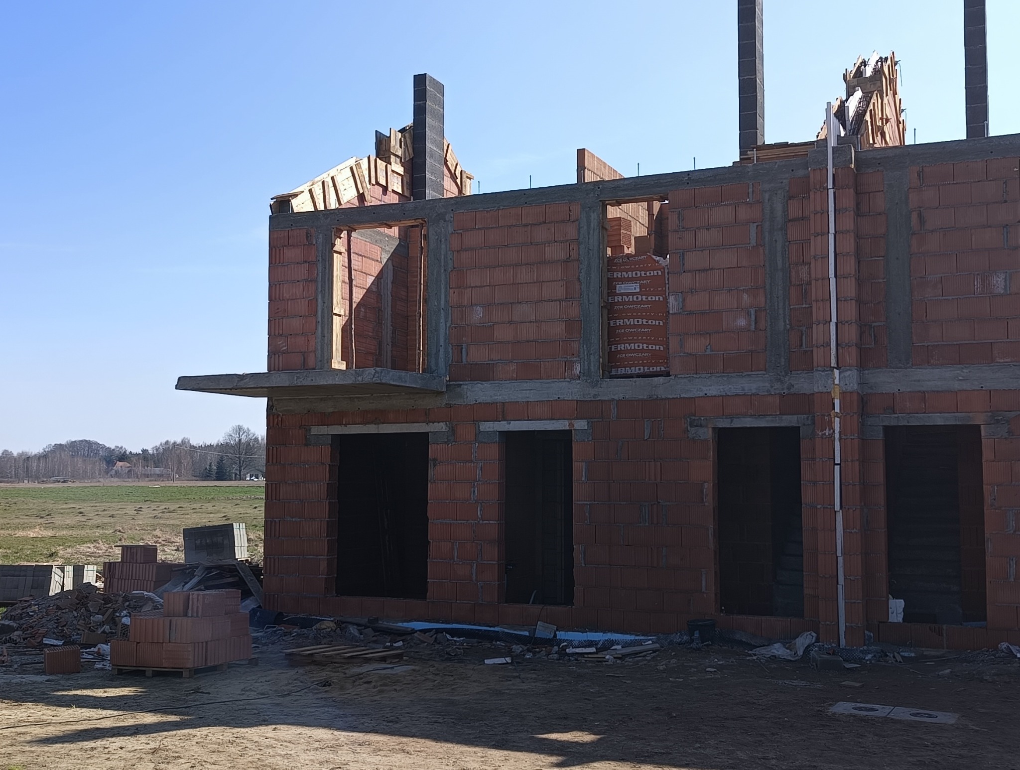 Wznoszenie dwukondygnacyjnego budynku z czerwonej cegły, widoczne otwory okienne i drzwiowe, częściowo wykonana konstrukcja dachu, palety cegieł obok budynku na tle łąki i błękitnego nieba.