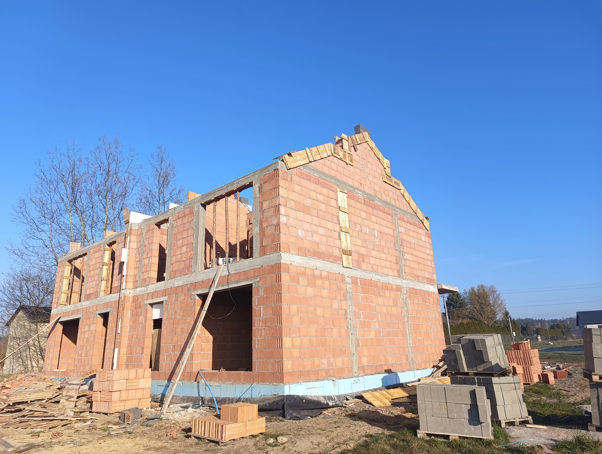 Surowy stan dwupiętrowego domu z czerwonej cegły, widoczne otwory okienne i drzwiowe, drewniane elementy szczytu dachu, palety z bloczkami obok.