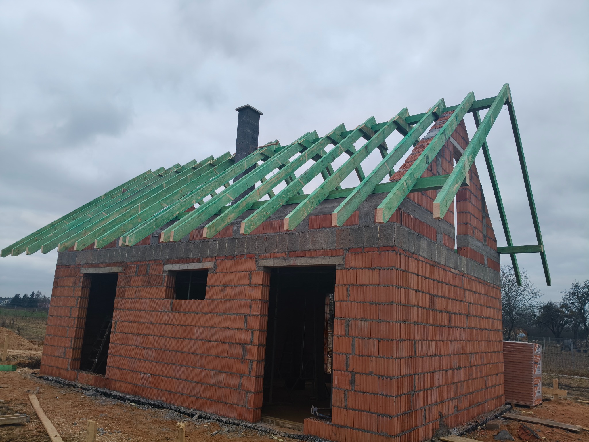 Niedokończony dom z czerwonej cegły z zieloną konstrukcją dachu i czarnym kominem na tle pochmurnego nieba. Widoczne otwarte przestrzenie na okna i drzwi.