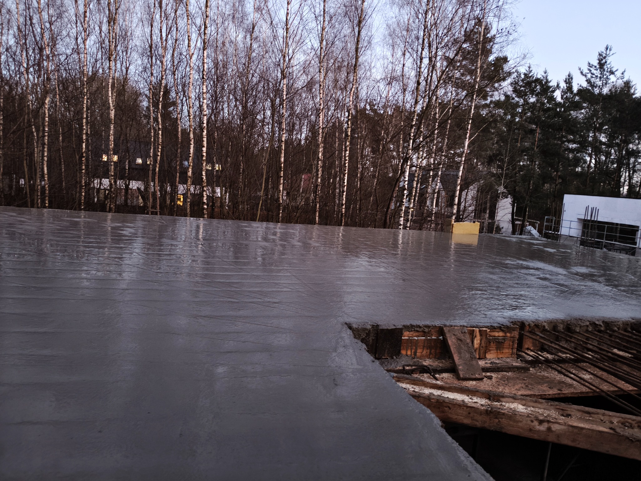 Świeżo wylany beton na stropie budynku w trakcie budowy, widoczne drewniane szalunki i zbrojenie. W tle drzewa bez liści i fragment budynku.