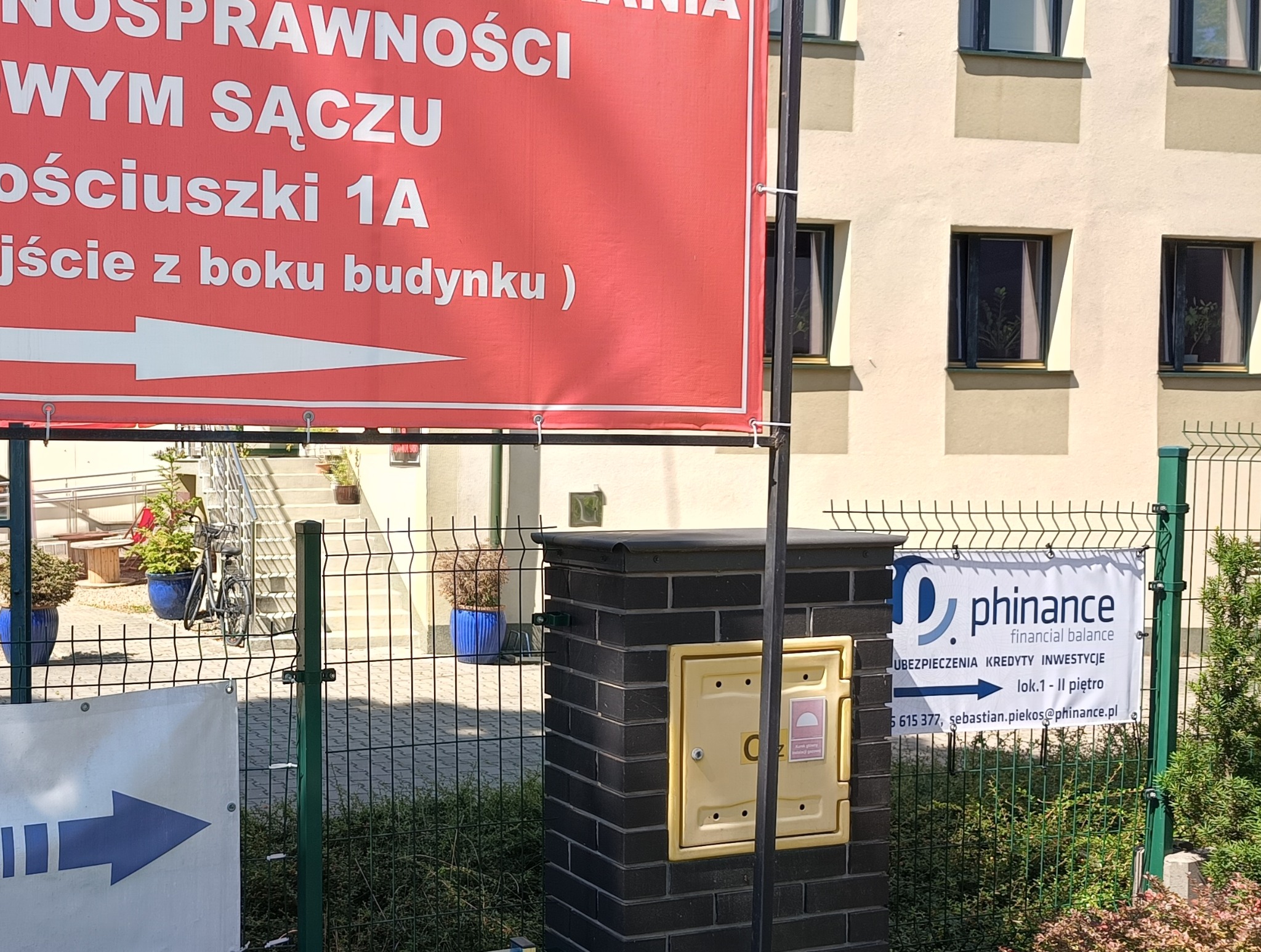 Baner reklamowy firmy Phinance z ofertą ubezpieczeń, kredytów i inwestycji, umieszczony na ogrodzeniu, obok skrzynki gazowej, w tle budynek i schody.