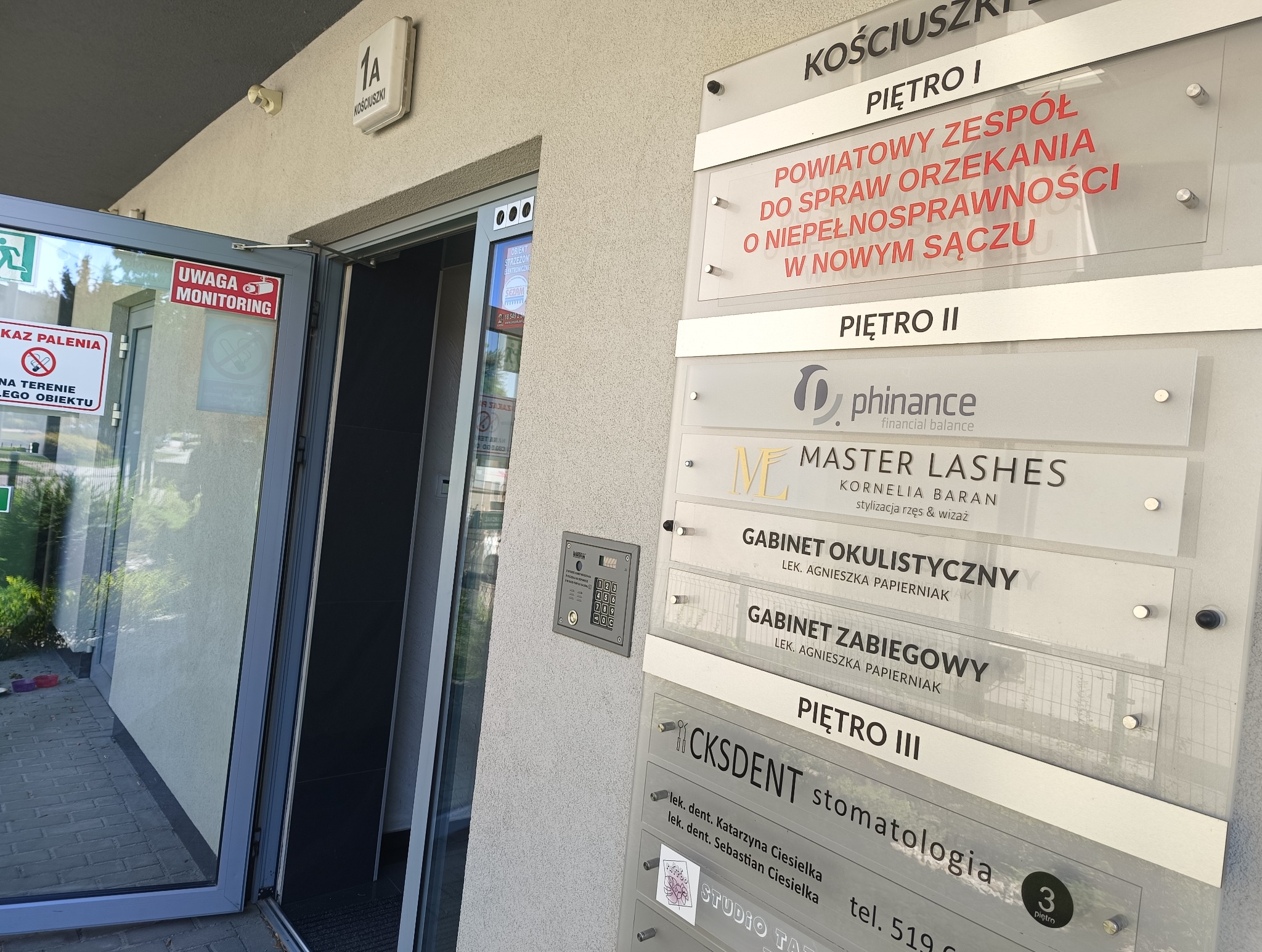 Tablica informacyjna przy wejściu do budynku z nazwami firm i gabinetów, w tym logo firmy 'phinance financial balance' na drugim piętrze.