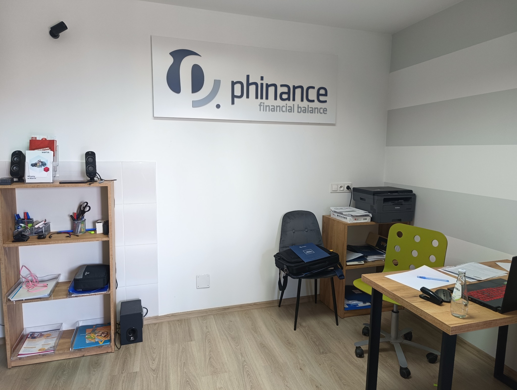 Wnętrze biura firmy Phinance z logo na ścianie, drewnianym regałem z dokumentami, biurkiem z laptopem i butelką wody.