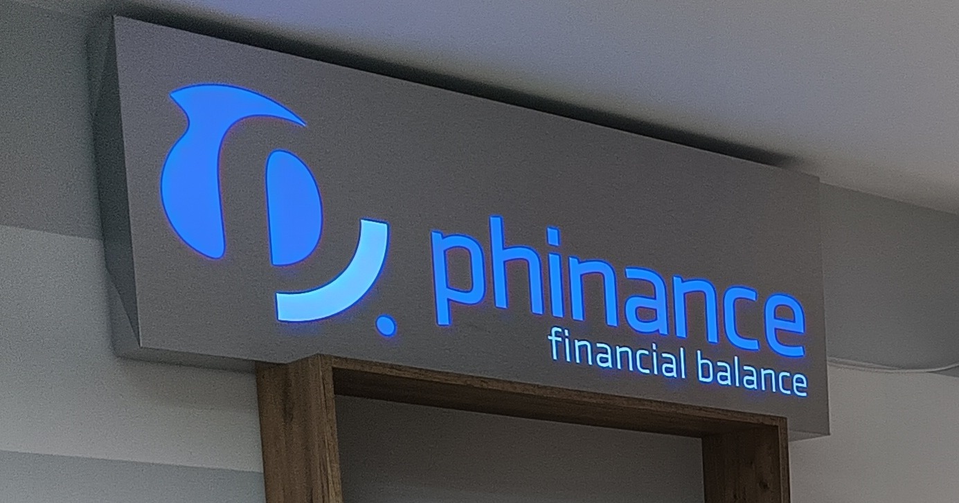 Podświetlany szyld firmy 'phinance financial balance' z niebieskim logo i nazwą, zamontowany na ścianie.