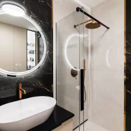 Elegancka łazienka z okrągłym lustrem LED, białą umywalką nablatową, kabiną prysznicową ze złotą armaturą i płytkami imitującymi marmur w odcieniach bieli i czerni.