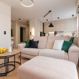 Royal Design Projektowanie Wnętrz Joanna Kowalska - Jasny salon z beżową sofą, pufem i okrągłym stolikiem kawowym. W tle widoczna kuchnia z beżowymi szafkami i nowoczesnym oświetleniem. Minimalistyczny design.