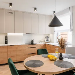 Jasna kuchnia z drewnianymi szafkami, okrągłym stołem z zielonymi krzesłami i czarną lampą stożkową. Na blacie czajnik i ekspres do kawy.