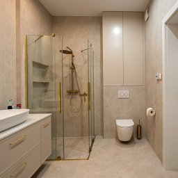 Royal Design Projektowanie Wnętrz Joanna Kowalska - Beżowa łazienka z kabiną prysznicową ze złotymi detalami, umywalką na szafce i toaletą. Minimalistyczny design, neutralna kolorystyka, oświetlenie punktowe.