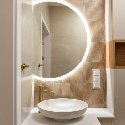 Royal Design Projektowanie Wnętrz Joanna Kowalska - Elegancka łazienka z okrągłym lustrem LED, umywalką nablatową i złotą armaturą. Dekoracyjna ściana z motywem jodełki. Subtelne oświetlenie i stonowana kolorystyka.