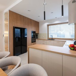 Royal Design Projektowanie Wnętrz Joanna Kowalska - Nowoczesna kuchnia z drewnianymi szafkami, czarną lodówką i wyspą z owocami. Minimalistyczne oświetlenie i jasne kolory tworzą przytulną atmosferę.