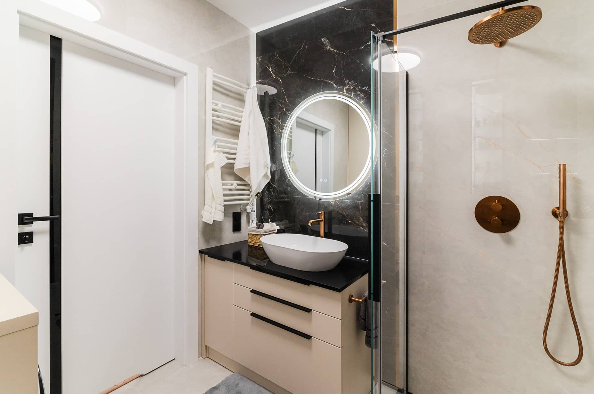 Nowoczesna łazienka z umywalką nablatową, okrągłym lustrem z podświetleniem LED, kabiną prysznicową i elementami w kolorze miedzi. Minimalistyczny design.