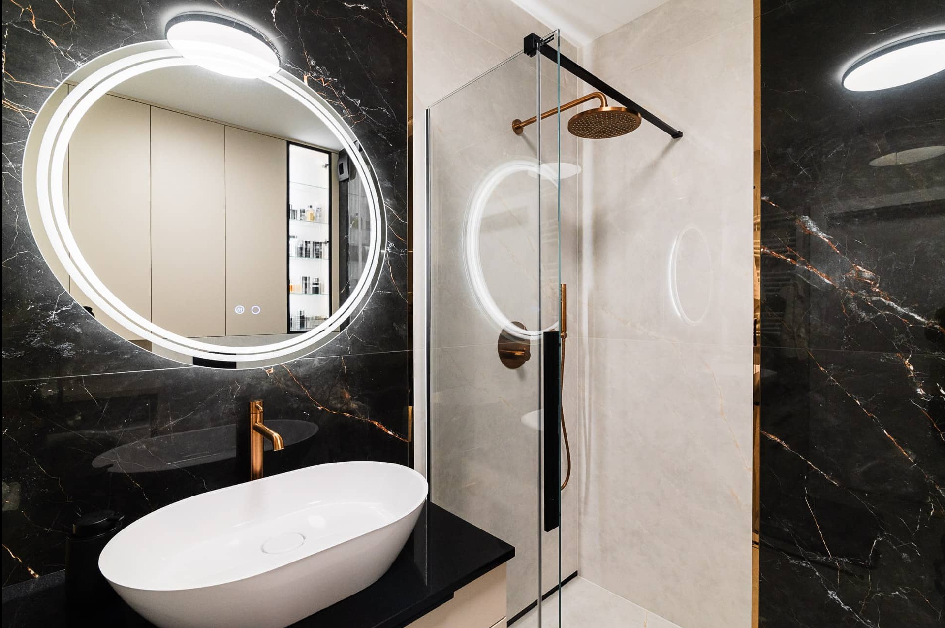 Elegancka łazienka z okrągłym lustrem LED, białą umywalką nablatową, kabiną prysznicową ze złotą armaturą i płytkami imitującymi marmur w odcieniach bieli i czerni.
