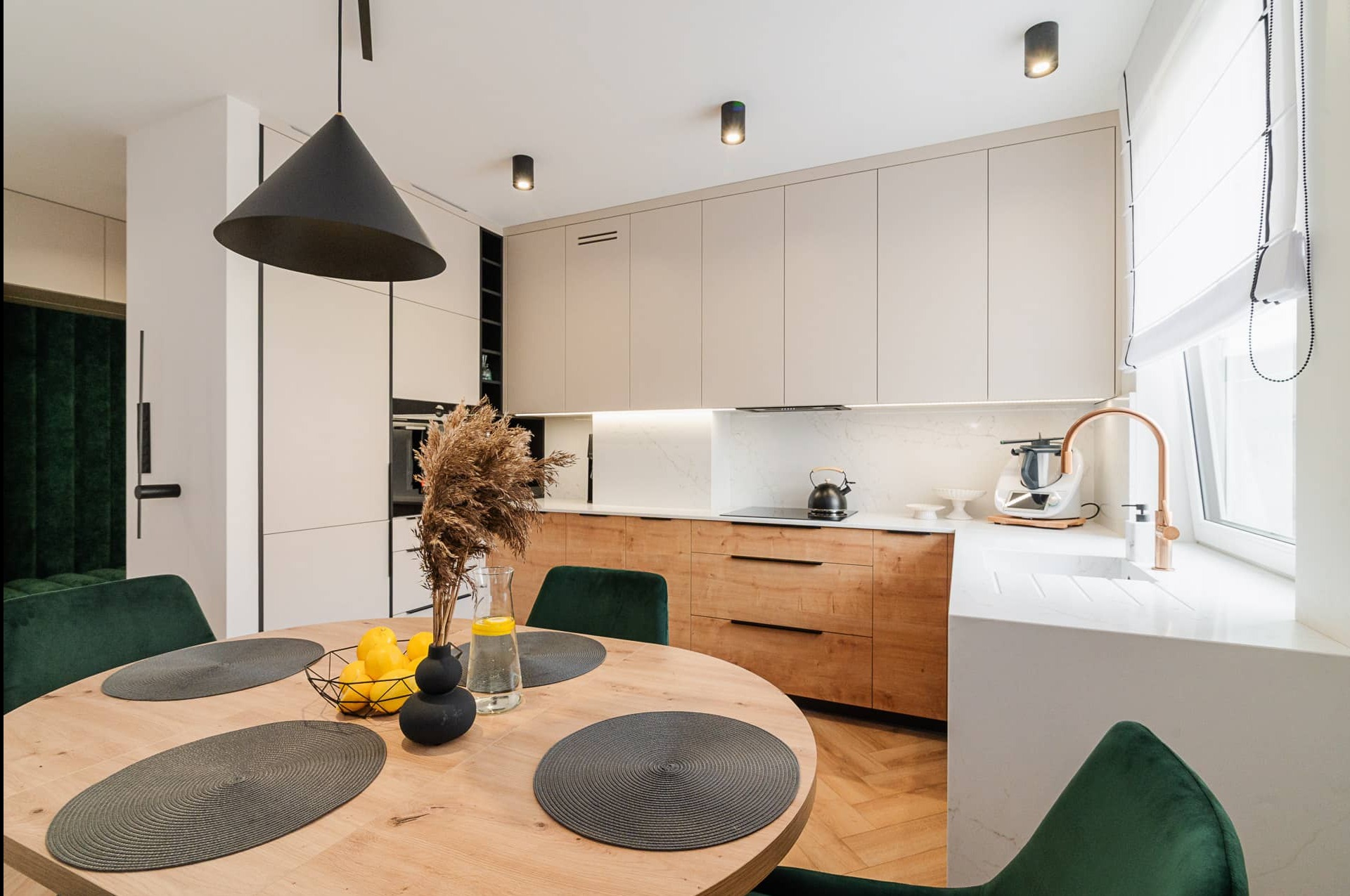Stylowa kuchnia z drewnianymi i beżowymi szafkami, okrągłym stołem z zielonymi krzesłami i czarną lampą stożkową. Nowoczesne oświetlenie punktowe.