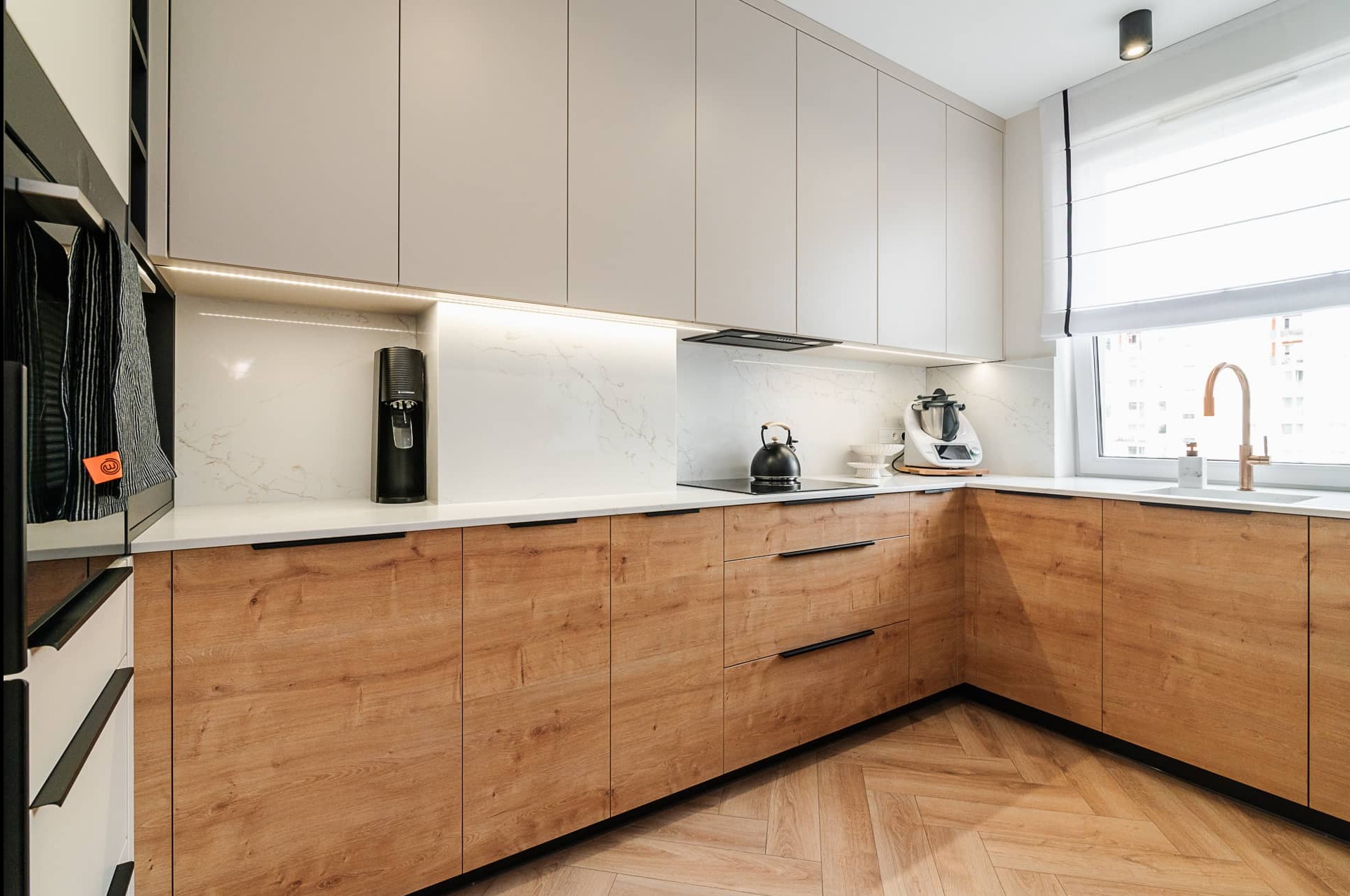 Nowoczesna kuchnia w beżach i drewnie: szafki z jasnego drewna, marmurowy backsplash, złota armatura i parkiet w jodełkę. Minimalistyczny design z akcentami luksusu.