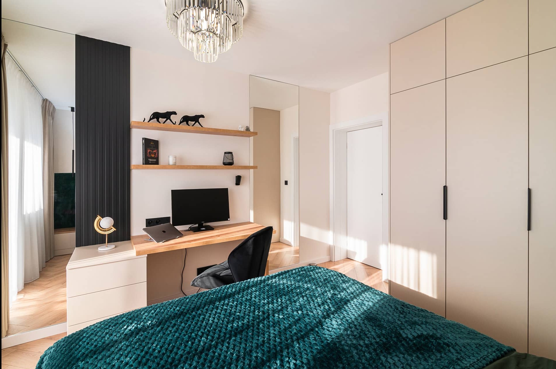 Nowoczesna sypialnia z biurkiem i szafą w zabudowie, oświetlona kryształowym żyrandolem. Na półkach dekoracyjne figurki panter i lampka na biurku.