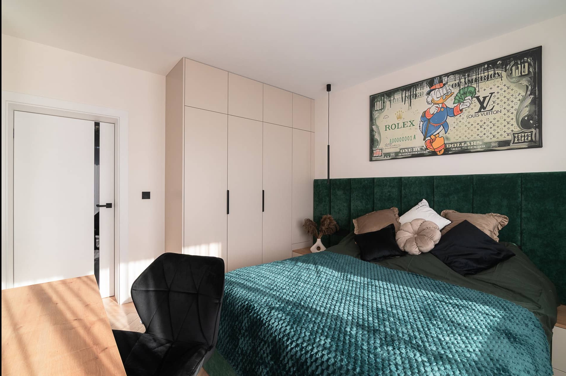 Sypialnia z zielonym zagłówkiem, turkusową narzutą i szafą w zabudowie. Na ścianie obraz z motywem banknotu i Kaczora Donalda, czarne krzesło obok biurka.