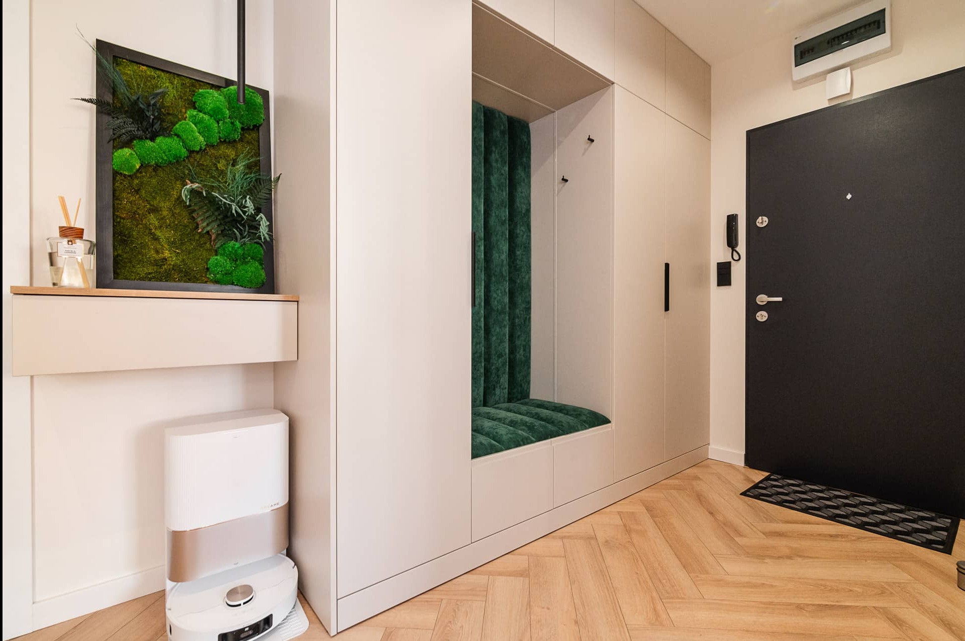 Nowoczesny przedpokój z siedziskiem w zabudowie, dekoracją z mchu i robotem sprzątającym. Podłoga w jodełkę, minimalistyczne wykończenie, czarne drzwi wejściowe.