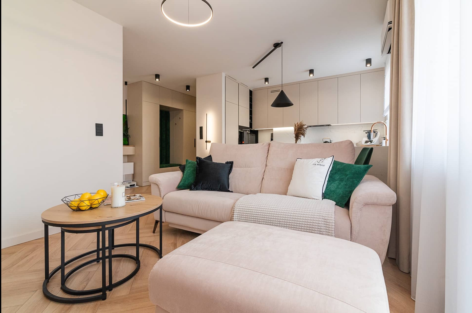 Jasny salon z beżową sofą, pufem i okrągłym stolikiem kawowym. W tle widoczna kuchnia z beżowymi szafkami i nowoczesnym oświetleniem. Minimalistyczny design.