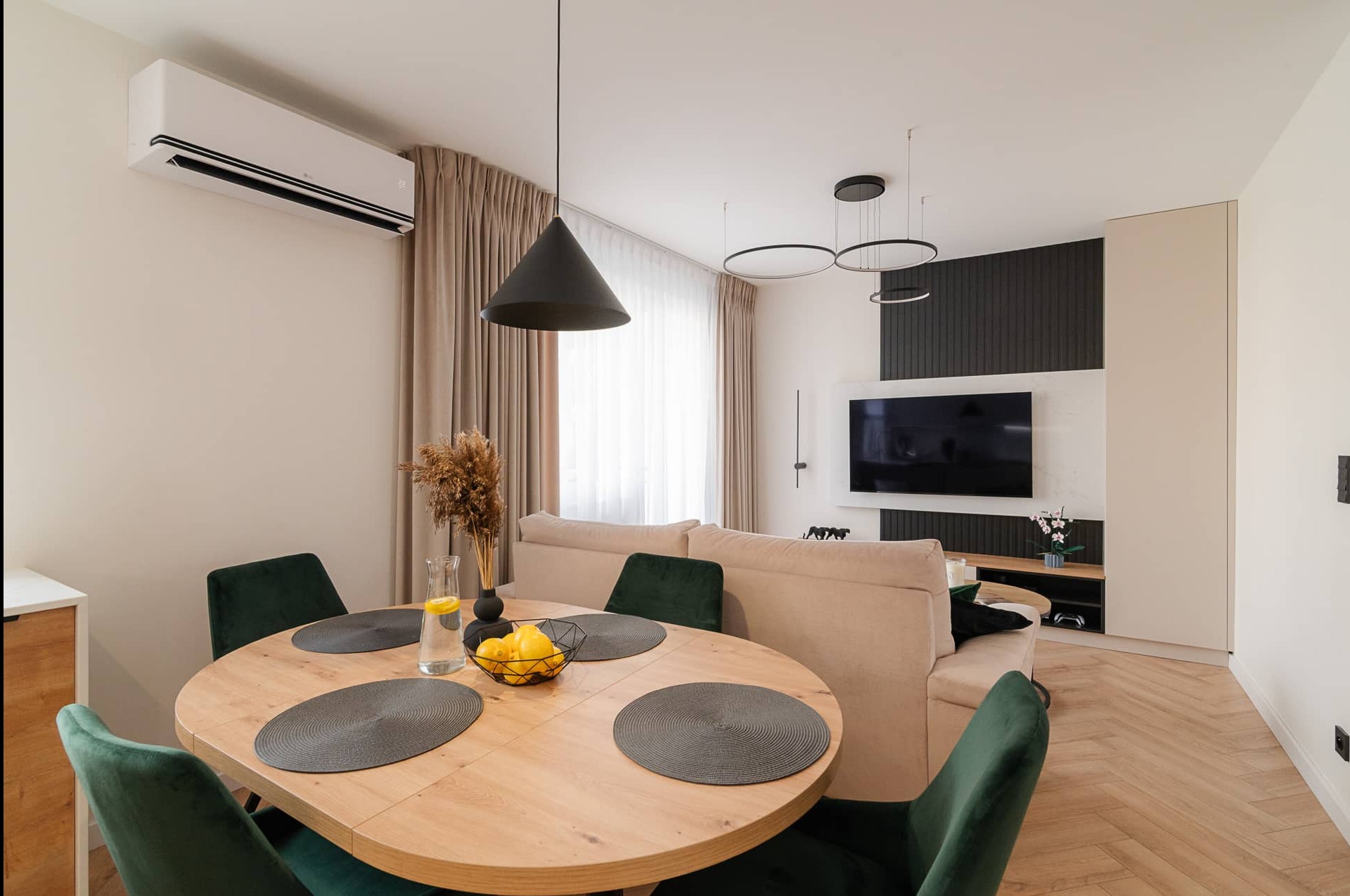 Nowoczesny salon z okrągłym stołem, zielonymi krzesłami, beżową sofą i telewizorem na tle ciemnej ściany. Minimalistyczne oświetlenie i parkiet w jodełkę.