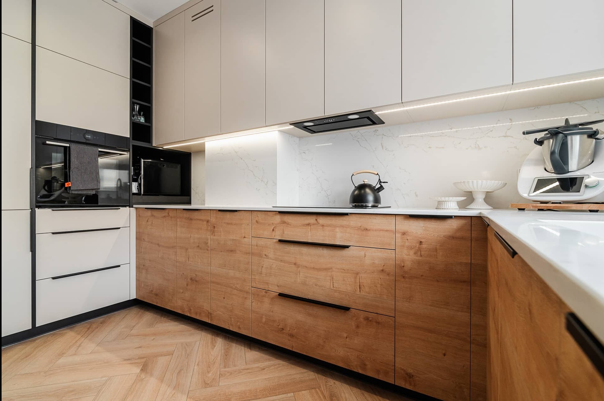 Nowoczesna kuchnia z drewnianymi frontami szafek i białym blatem, oświetlona listwą LED, z czajnikiem i robotem kuchennym na blacie. Parkiet w jodełkę.
