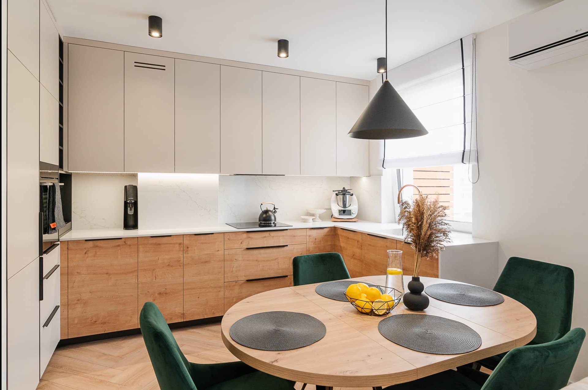 Jasna kuchnia z drewnianymi szafkami, okrągłym stołem z zielonymi krzesłami i czarną lampą stożkową. Na blacie czajnik i ekspres do kawy.