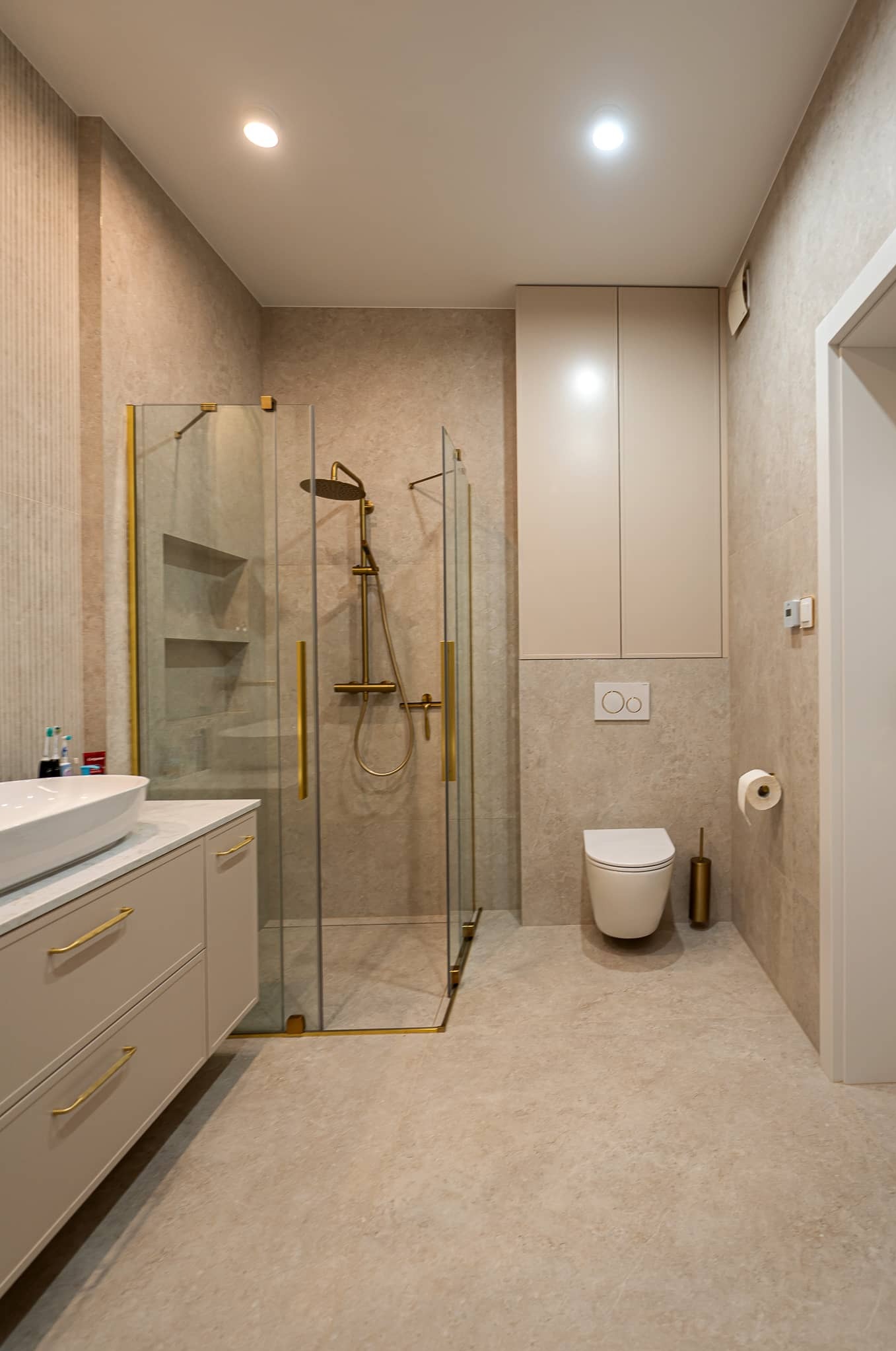 Beżowa łazienka z kabiną prysznicową ze złotymi detalami, umywalką na szafce i toaletą. Minimalistyczny design, neutralna kolorystyka, oświetlenie punktowe.