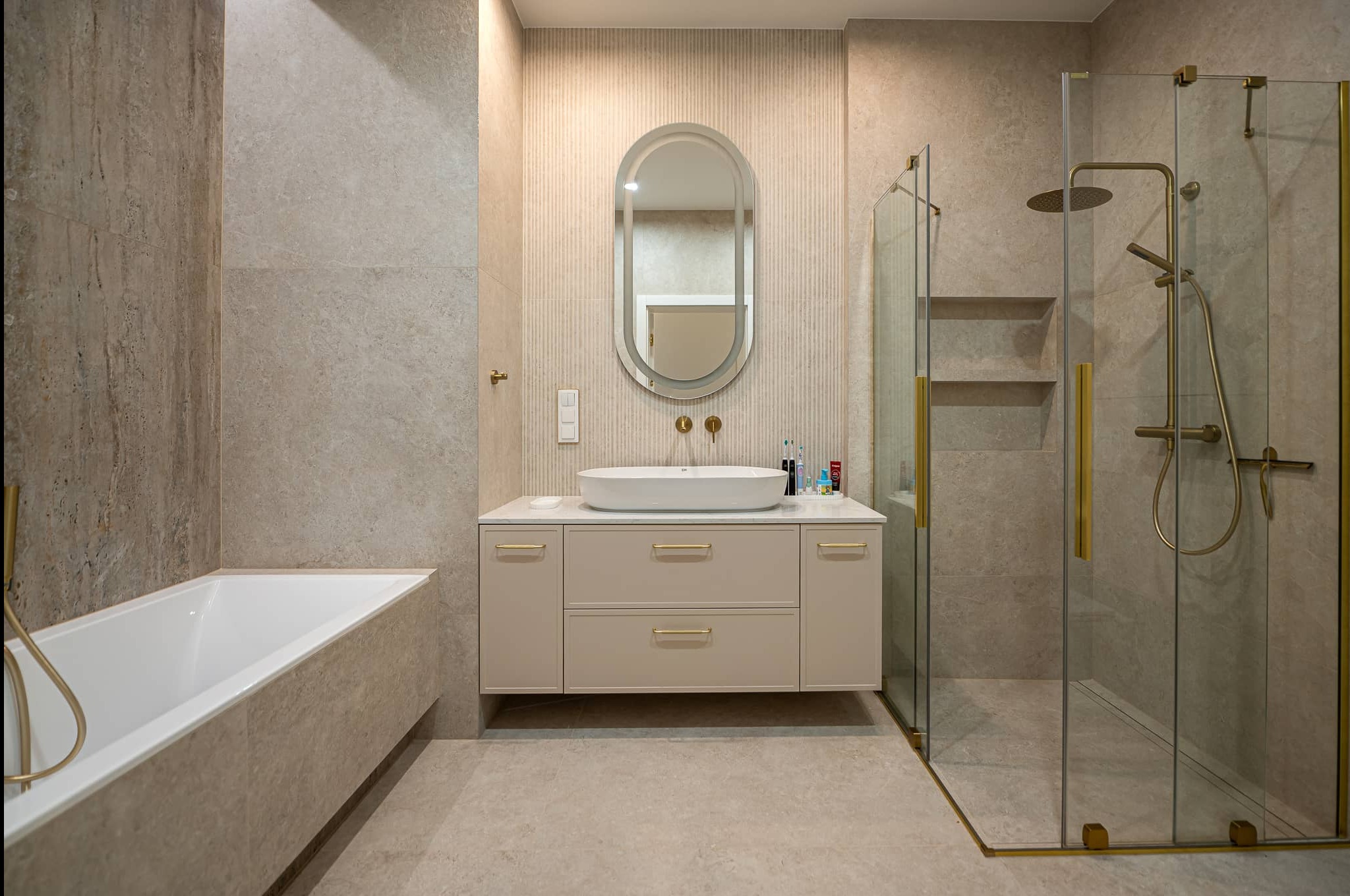 Elegancka łazienka w beżach: owalne lustro z podświetleniem, biała umywalka nablatowa, kabina prysznicowa ze złotymi detalami i wanna.