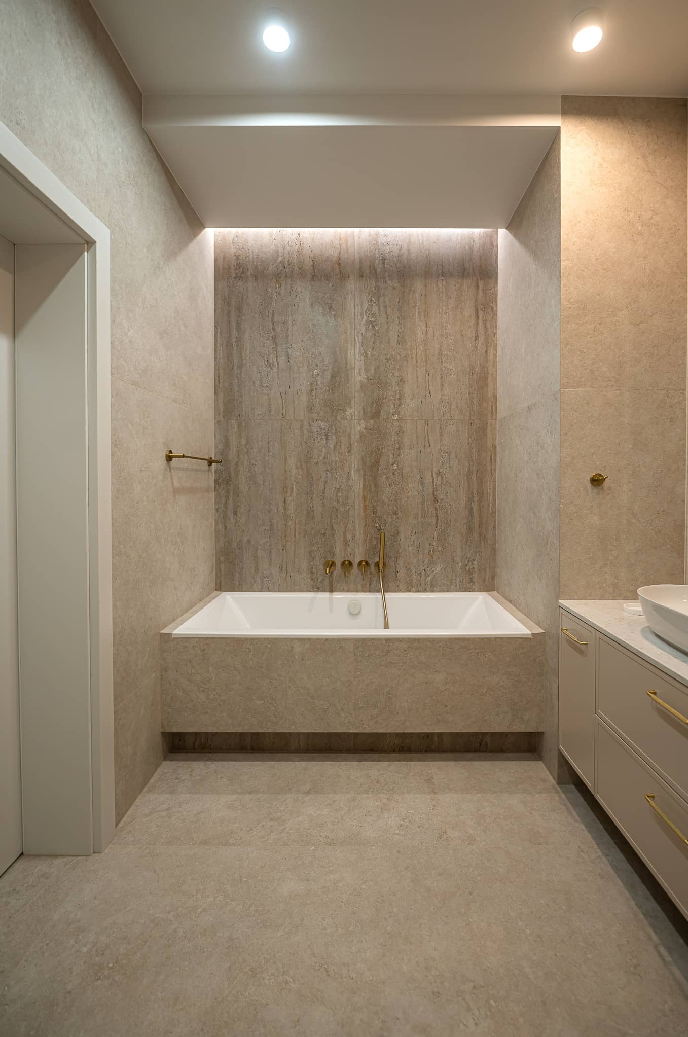 Elegancka łazienka w beżach: wanna obudowana płytkami, złota armatura, oświetlenie LED. Minimalistyczny design z dbałością o detale i spójną kolorystykę.