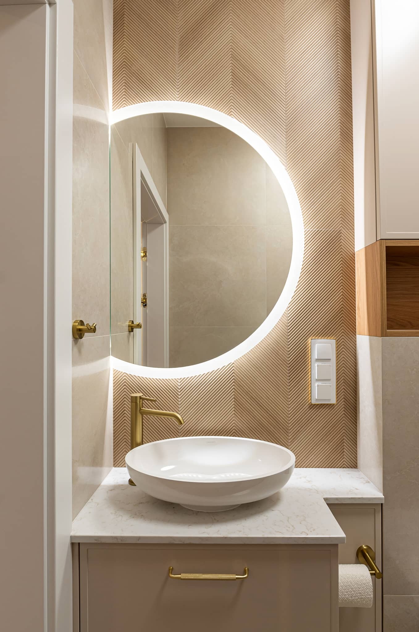 Elegancka łazienka z okrągłym lustrem LED, umywalką nablatową i złotą armaturą. Dekoracyjna ściana z motywem jodełki. Subtelne oświetlenie i stonowana kolorystyka.