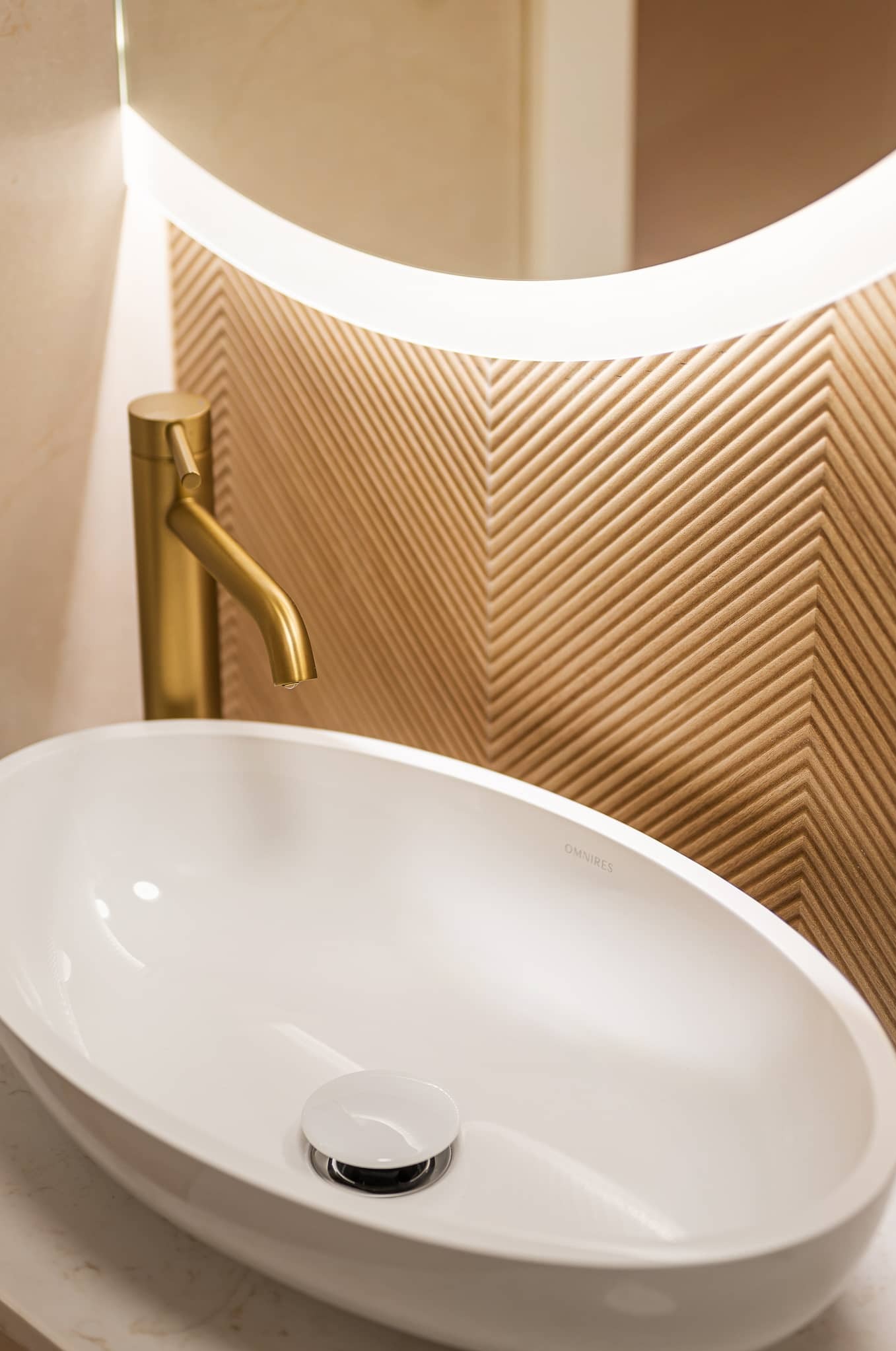 Nowoczesna łazienka: biała umywalka OMNIRES, złota bateria, podświetlane lustro i dekoracyjna ściana z drewnianych paneli ułożonych w jodełkę.