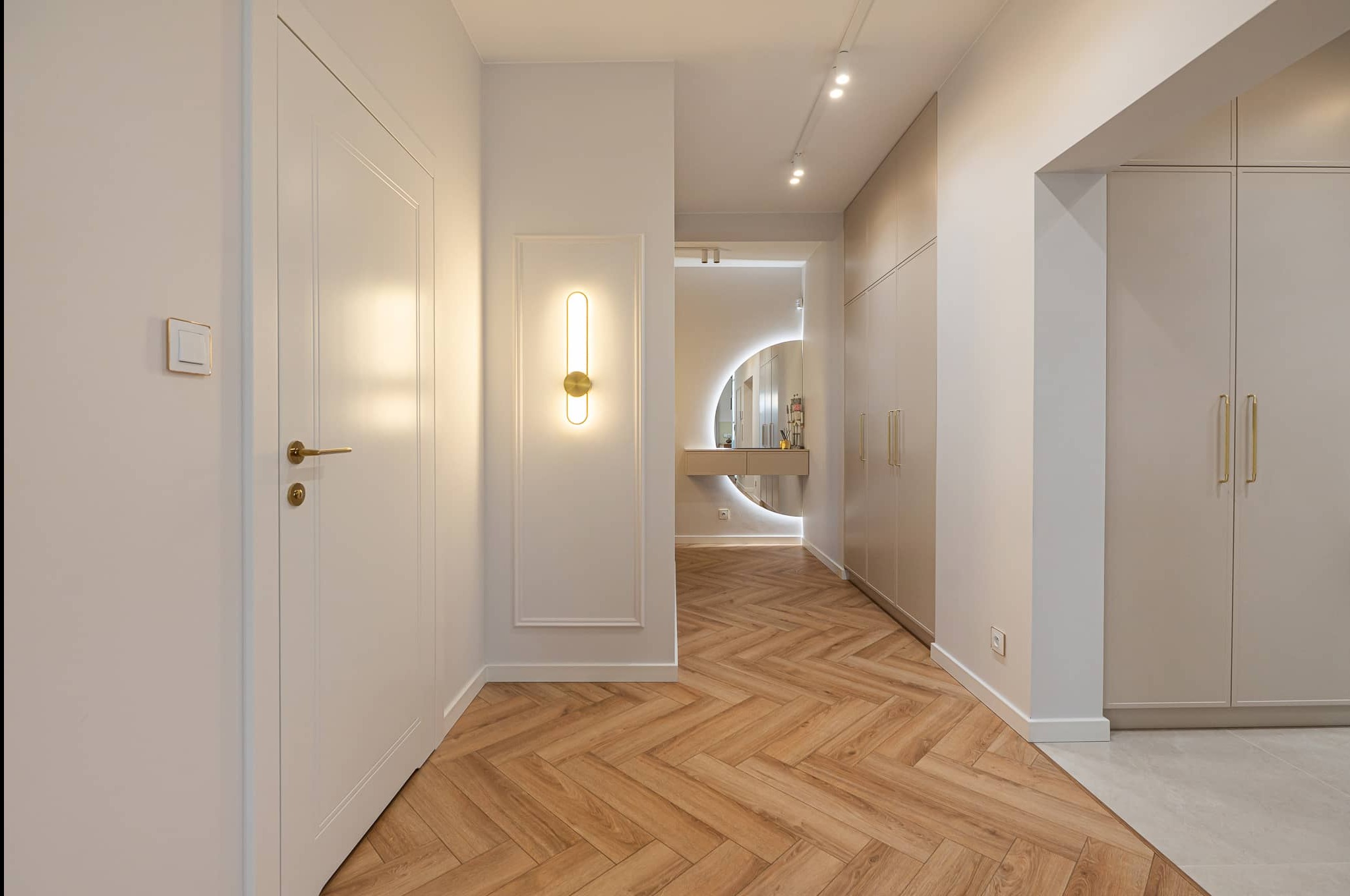 Korytarz w jasnych barwach z parkietem w jodełkę. Białe drzwi z złotą klamką, oświetlenie punktowe i dekoracyjna lampa ścienna. W tle lustro z podświetleniem.