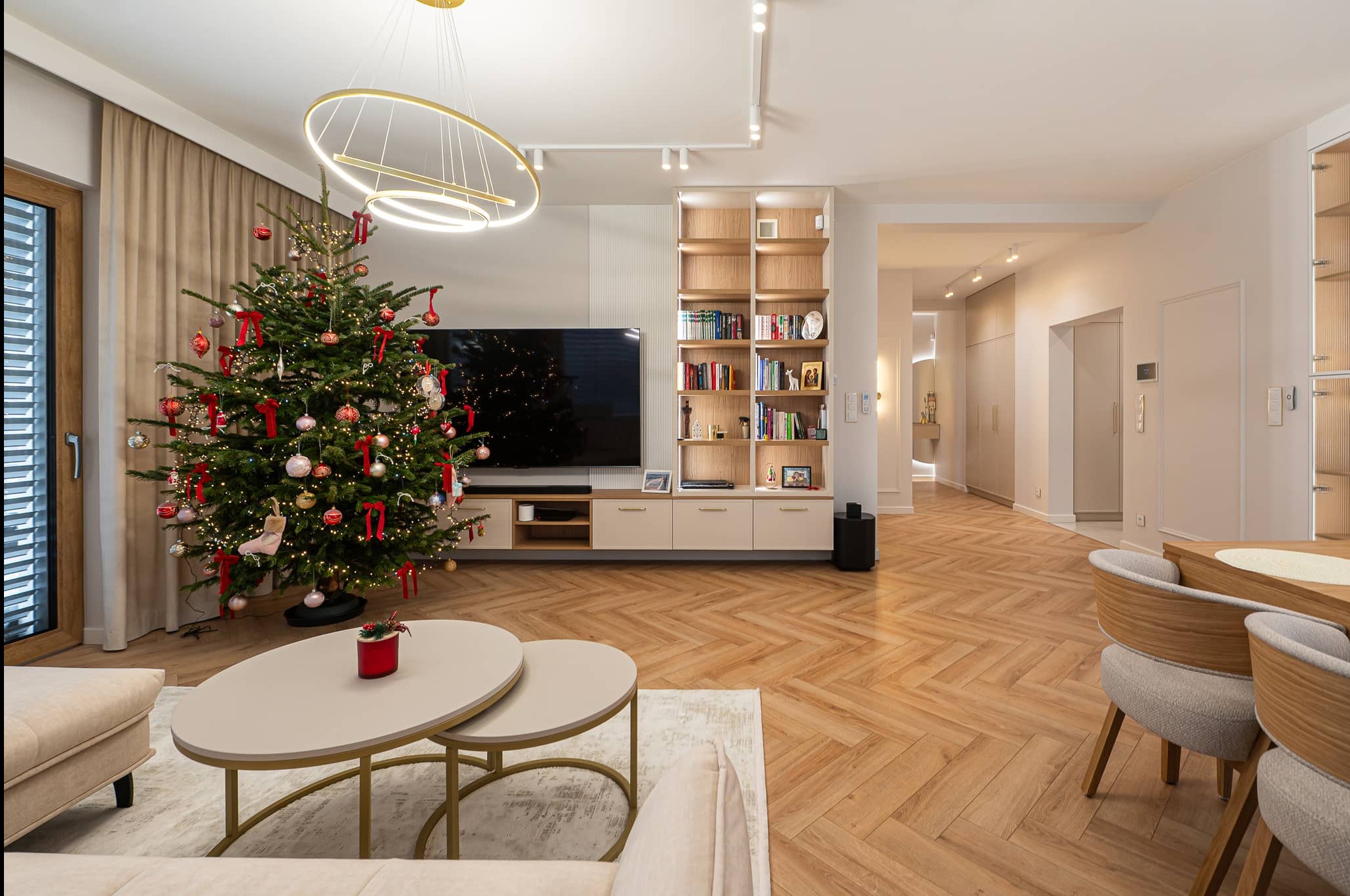 Świątecznie udekorowany salon z parkietem w jodełkę, choinką, nowoczesną lampą i regałem z książkami. Jasne, przestronne wnętrze z elementami dekoracyjnymi.