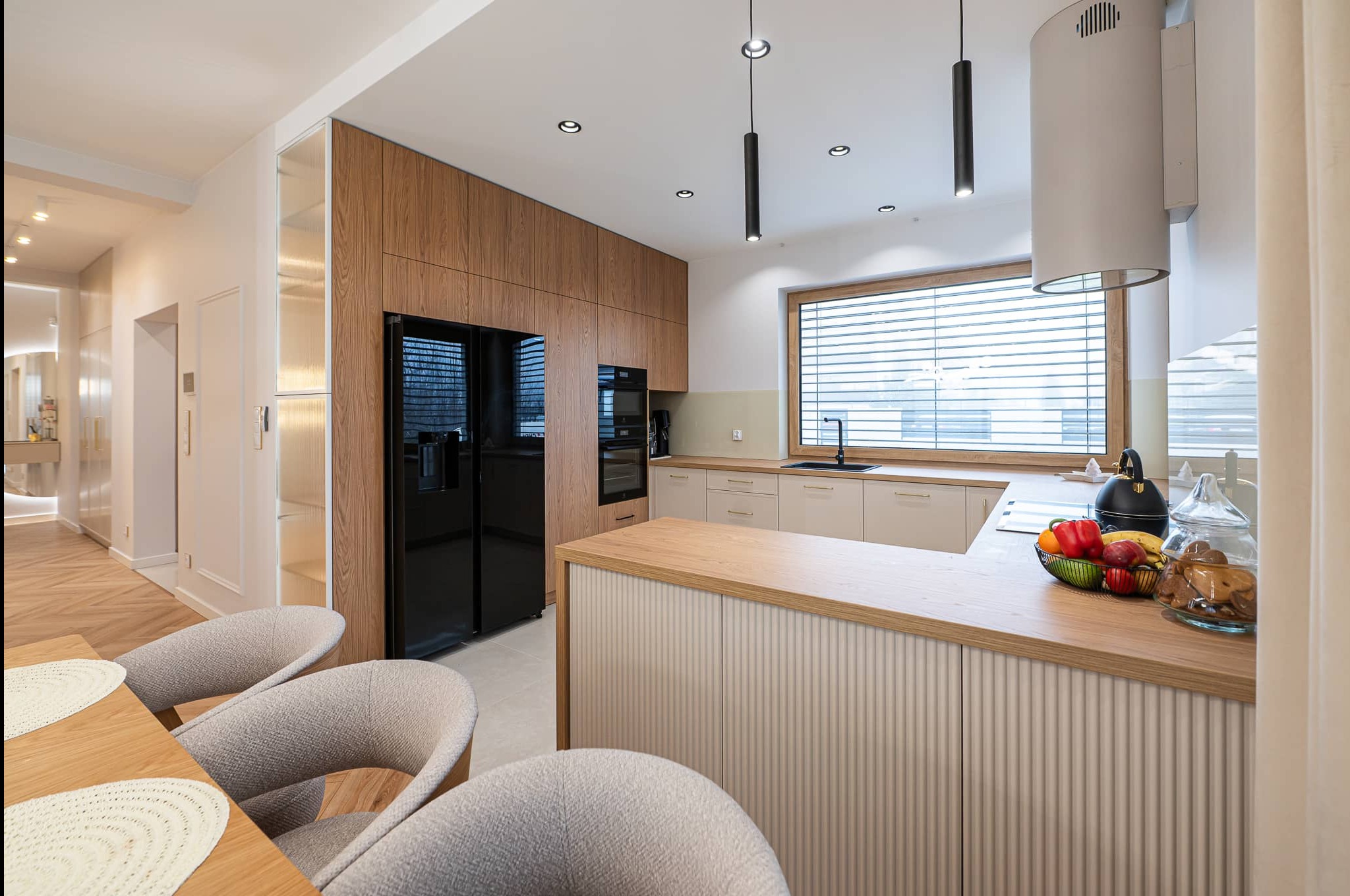 Nowoczesna kuchnia z drewnianymi szafkami, czarną lodówką i wyspą z owocami. Minimalistyczne oświetlenie i jasne kolory tworzą przytulną atmosferę.